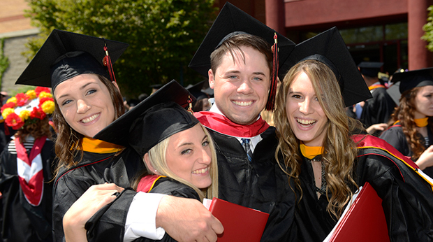 Four La Roche University graduates wearing their caps and gowns celebrate after commencement. They all smile, and two of them have their arms around each other. 